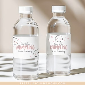 May include: Two clear glass water bottles with white labels. The labels have a pink cartoon dumpling with the text "Our little dumpling is on the way".