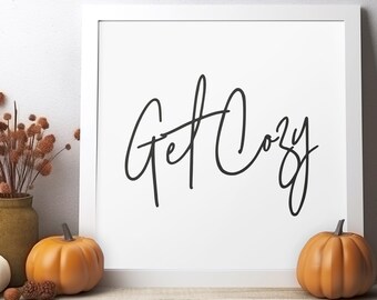 May include: White framed print with black calligraphy that reads "Get Cozy". The print is sitting on a wooden surface with two orange pumpkins and a white pumpkin in front of it.