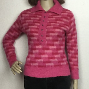 May include: A pink sweater with a collared neckline and a button-down placket. The sweater features a geometric pattern in shades of pink and is paired with black pants. The mannequin is wearing a short brown wig.