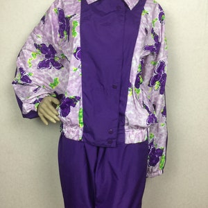May include: A vintage purple and floral print tracksuit. The jacket features a purple center panel with snap closures and a floral print on the sleeves and shoulders. The matching pants are solid purple.