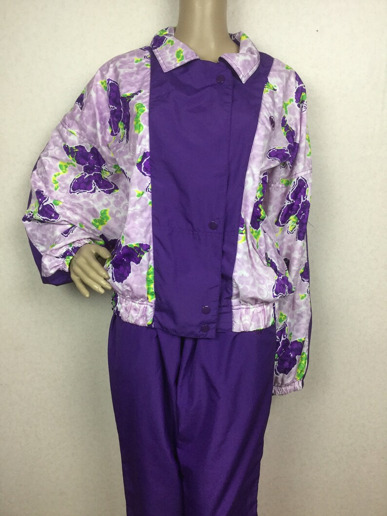 May include: A vintage purple and floral print tracksuit. The jacket features a purple center panel with snap closures and a floral print on the sleeves and shoulders. The matching pants are solid purple.