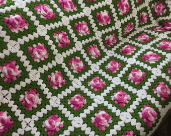 May include: A handmade crochet blanket featuring a pattern of pink roses on a green and white background. The blanket has a textured appearance with the raised floral details. The edges are finished with a white fringe.