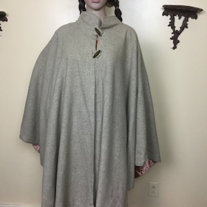 May include: A beige cape featuring a subtle plaid pattern, a high collar, and two brown toggle closures at the neck. The garment has a loose, flowing silhouette and is lined with a smooth, pink fabric visible at the bottom edges of the sleeves.