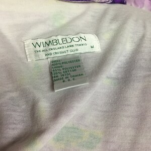 May include: Close-up of a vintage Wimbledon tennis jacket. The jacket features a white interior with a floral pattern and a purple collar. The Wimbledon label is visible, along with the fabric composition and size M.