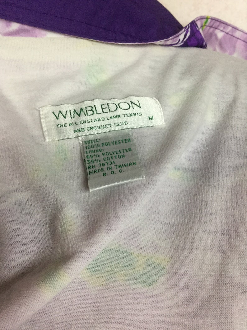 May include: Close-up of a vintage Wimbledon tennis jacket. The jacket features a white interior with a floral pattern and a purple collar. The Wimbledon label is visible, along with the fabric composition and size M.