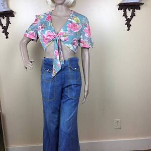 May include: A blue and pink floral print crop top with a tie front and short sleeves. The top is paired with blue denim bell bottom pants with yellow stitching.