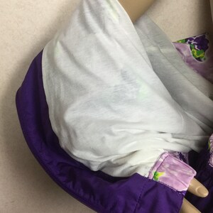 May include: A close-up of a purple and white jacket with a hood. The hood is lined with white fabric and trimmed with purple. The jacket has a floral pattern on the sleeve and is zipped up.