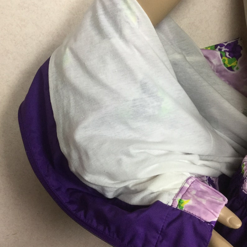 May include: A close-up of a purple and white jacket with a hood. The hood is lined with white fabric and trimmed with purple. The jacket has a floral pattern on the sleeve and is zipped up.