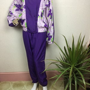 May include: A vintage purple and floral print tracksuit. The jacket features a purple center panel and floral print sleeves and upper body. The matching pants are solid purple. The floral print includes purple flowers with green accents on a light purple background.
