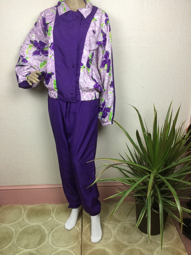 May include: A vintage purple and floral print tracksuit. The jacket features a purple center panel and floral print sleeves and upper body. The matching pants are solid purple. The floral print includes purple flowers with green accents on a light purple background.