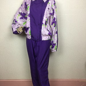 May include: A vintage purple tracksuit set. The jacket features a floral pattern in shades of purple and green on a light pink background, with a solid purple center panel. The matching pants are a solid purple color.