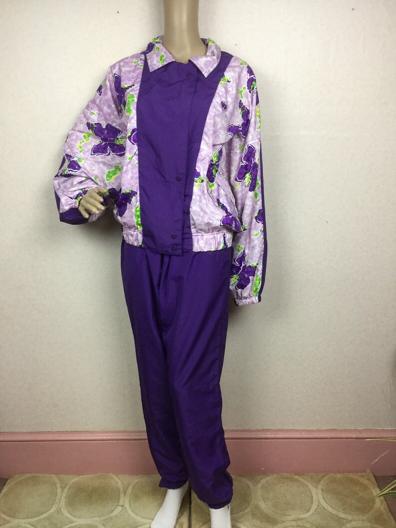 May include: A vintage purple tracksuit set. The jacket features a floral pattern in shades of purple and green on a light pink background, with a solid purple center panel. The matching pants are a solid purple color.