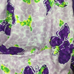 May include: A close-up of a fabric with a repeating butterfly pattern. The butterflies are purple with green accents, set against a light purple background with a speckled design. The fabric is folded, showing the pattern's details.