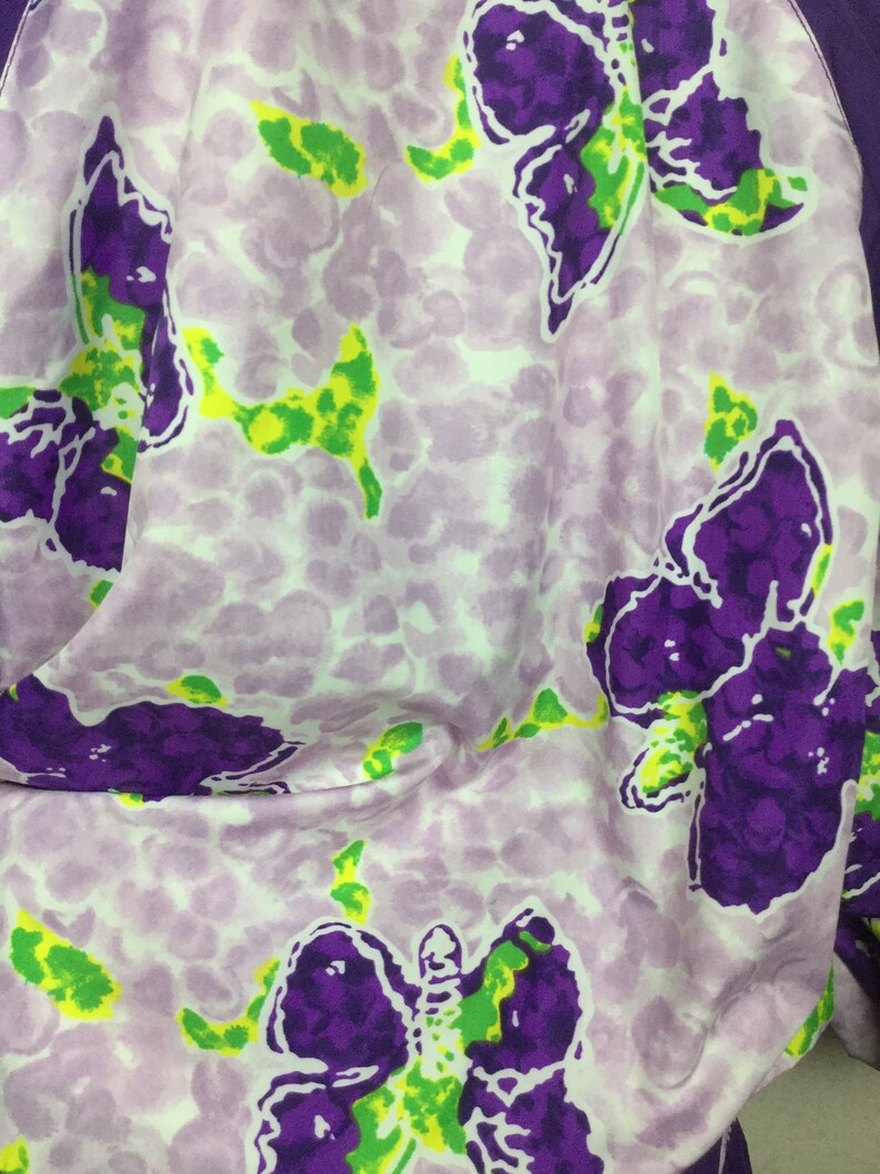 May include: A close-up of a fabric with a repeating butterfly pattern. The butterflies are purple with green accents, set against a light purple background with a speckled design. The fabric is folded, showing the pattern's details.