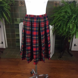 May include: A red, green, and blue plaid skirt with pleats. The skirt is on a white mannequin and is displayed on a wooden floor.