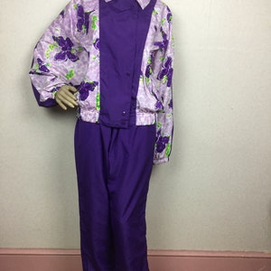 May include: A vintage purple tracksuit set. The jacket features a floral pattern in shades of purple and green on a light purple background, with a solid purple panel down the front. The matching pants are a solid, deep purple color. The jacket has a button closure.