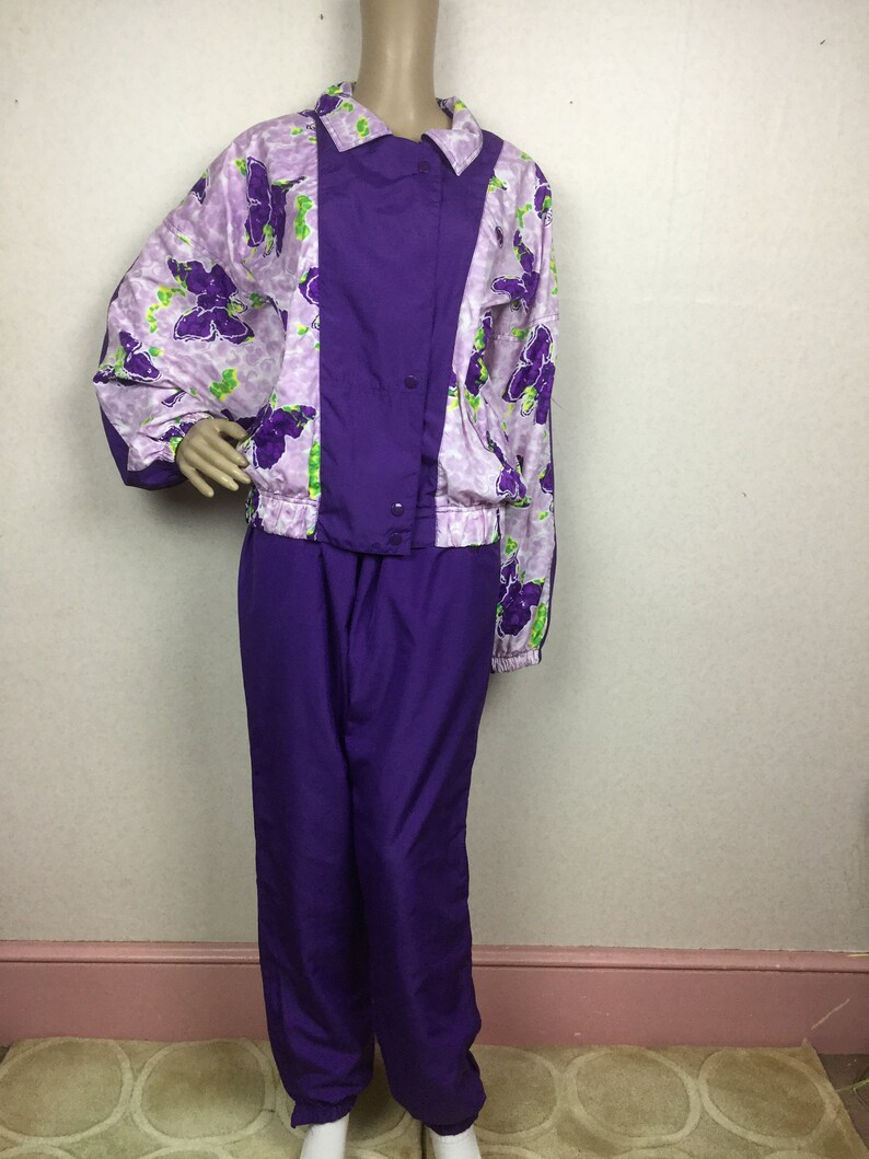 May include: A vintage purple tracksuit set. The jacket features a floral pattern in shades of purple and green on a light purple background, with a solid purple panel down the front. The matching pants are a solid, deep purple color. The jacket has a button closure.