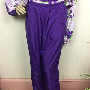 May include: A two-piece purple tracksuit. The pants are a solid, vibrant purple with an elastic waistband. The jacket is a light purple with a floral pattern in shades of purple and green. The outfit is displayed on a mannequin.