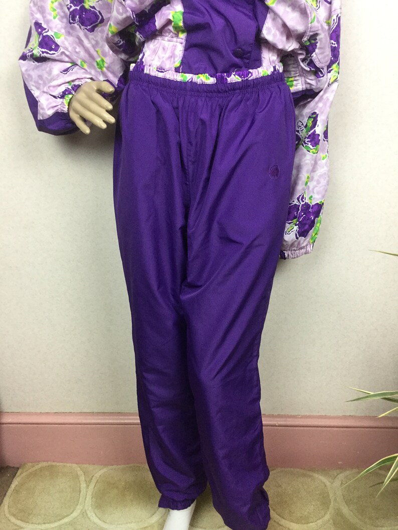 May include: A two-piece purple tracksuit. The pants are a solid, vibrant purple with an elastic waistband. The jacket is a light purple with a floral pattern in shades of purple and green. The outfit is displayed on a mannequin.