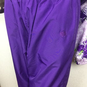 May include: Close-up of a pair of vibrant purple athletic pants with an elastic waistband. The pants feature a small, embroidered emblem on the left leg. The fabric has a slight sheen. Part of a floral patterned jacket is visible.