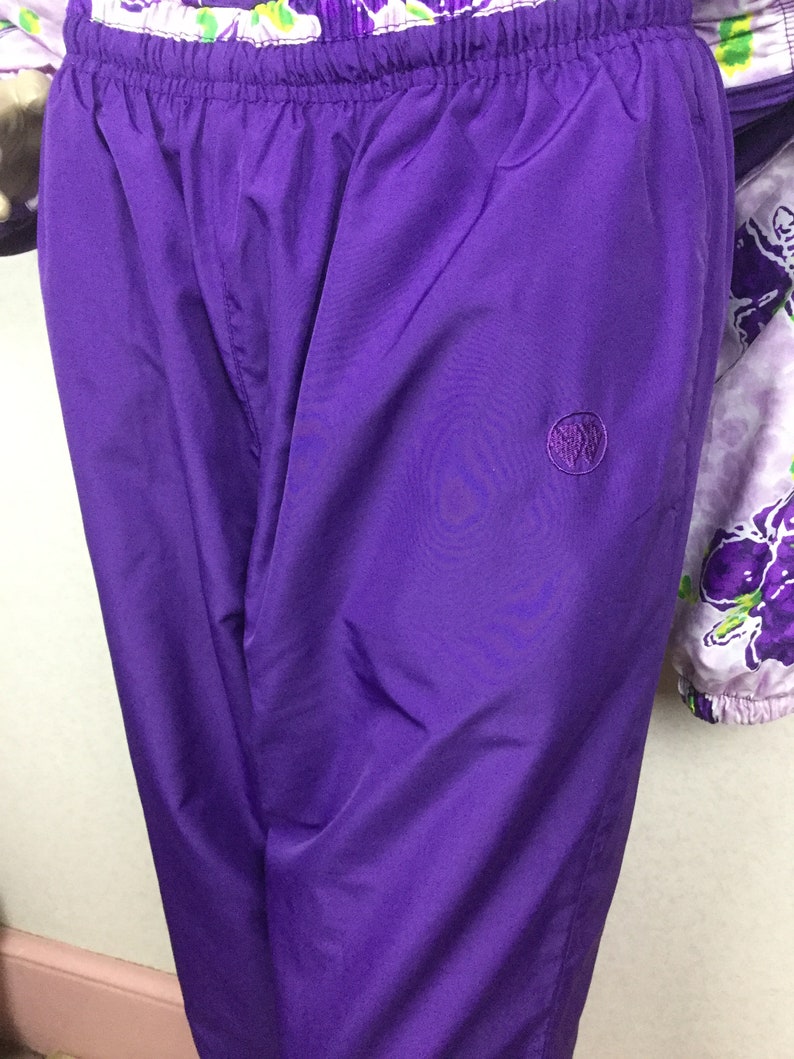 May include: Close-up of a pair of vibrant purple athletic pants with an elastic waistband. The pants feature a small, embroidered emblem on the left leg. The fabric has a slight sheen. Part of a floral patterned jacket is visible.