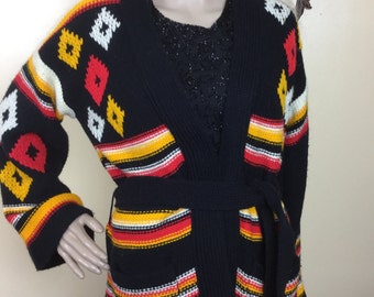 May include: A black knit cardigan with a geometric pattern in red, yellow, and white. The cardigan has a belt, pockets, and a shawl collar. The sleeves and body feature horizontal stripes in red, yellow, and white. The cardigan is displayed on a mannequin.