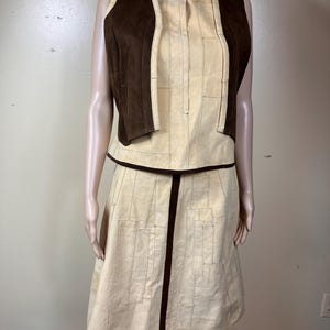 May include: A sleeveless vest and a knee-length skirt set in cream and dark brown. The vest features dark brown suede-like side panels with cream front panels. The skirt and vest front have a geometric stitched pattern with dark brown trim.
