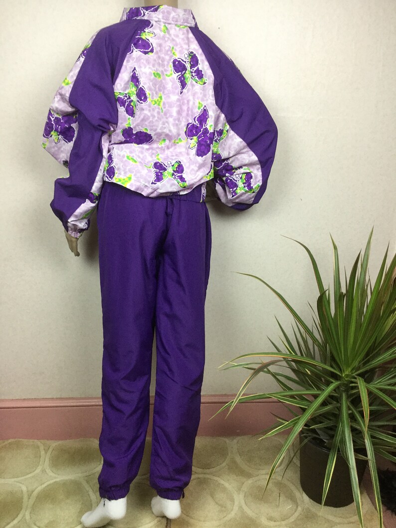 May include: A vintage purple tracksuit. The jacket features a butterfly print on a light purple background, with solid purple sleeves and collar. The matching pants are a solid purple color. The tracksuit is displayed on a mannequin.