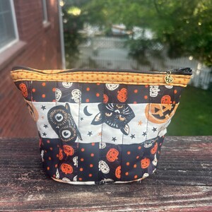 May include: A quilted fabric zipper pouch featuring a Halloween-themed pattern with black cats, owls, and orange pumpkins on a black and white background. The pouch has a yellow dotted trim along the top edge and a small gold heart-shaped zipper pull.