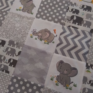 May include: A gray and white baby quilt with a patchwork design. The quilt features embroidered elephants in various poses, including a baby elephant holding a flower.
