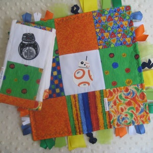 May include: A multi-colored baby quilt with a patchwork design featuring Star Wars characters BB-8 and a black and white droid. The quilt has colorful ribbons attached to the edges.