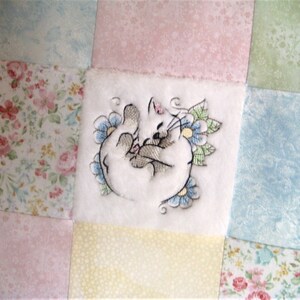May include: A white fabric square with a colorful embroidered cat design. The fabric is surrounded by squares of different floral patterned fabrics in pink, blue, and green.