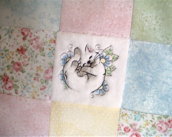 Puede incluir: Un cuadrado de tela blanca con un diseño de gato bordado de colores. La tela está rodeada de cuadrados de telas con diferentes patrones florales en rosa, azul y verde.