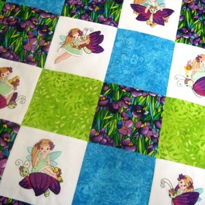 May include: A white quilt with a patchwork design featuring green, blue, and white squares. Each square has an embroidered image of a fairy with a butterfly and flowers.