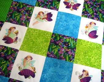 Puede incluir: Una colcha blanca con un diseño de patchwork con cuadrados verdes, azules y blancos. Cada cuadrado tiene una imagen bordada de un hada con una mariposa y flores.