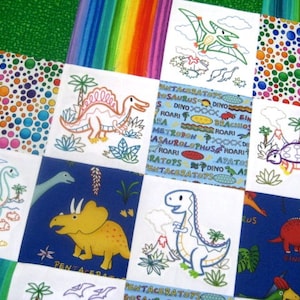 May include: A colorful quilt with a dinosaur theme. Each square features a different dinosaur, including a stegosaurus, triceratops, and a pterodactyl. The quilt is made of white fabric with colorful embroidered dinosaurs and text.