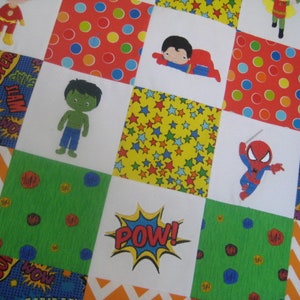 May include: A colorful quilt with a patchwork design featuring various superhero characters, including Superman, Hulk, Spiderman, and Flash. The quilt has a white background with squares of red, green, yellow, blue, and orange fabric. The squares are decorated with polka dots, stars, and comic book-style speech bubbles.