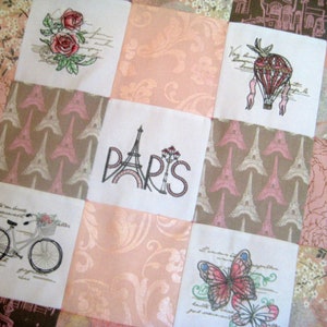 May include: A pink and brown patchwork quilt with embroidered images of a hot air balloon, Eiffel Tower, roses, a bicycle, and a butterfly. The quilt has the word "PARIS" embroidered in the center.