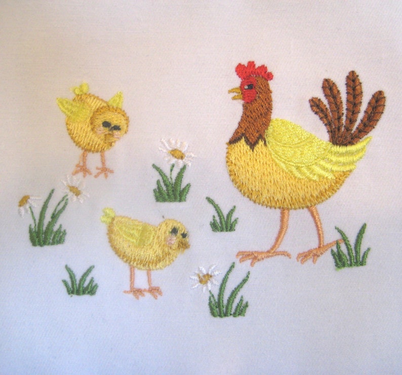 May include: Embroidered design of a yellow hen with a red comb and two yellow chicks. The hen is standing on a white background with green grass and white flowers.
