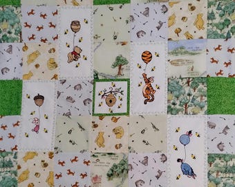 Puede incluir: Una colcha de retazos con varios paneles de tela cuadrados que muestran personajes de Winnie the Pooh, incluidos Pooh, Tigger, Piglet y Eeyore, cada uno sosteniendo un globo, intercalados con escenas de bosque, tarros de miel y una colmena sobre un borde verde.