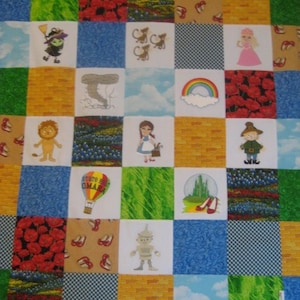 May include: A colorful patchwork quilt featuring characters and scenes from the Wizard of Oz. The quilt includes a yellow brick road, a tornado, a rainbow, Dorothy, the Wicked Witch of the West, the Cowardly Lion, the Tin Man, the Scarecrow, and the Emerald City.