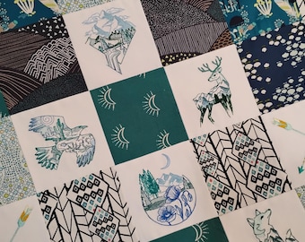 Puede incluir: Una colcha de retazos con una cuadrícula de cuadrados alternos. Algunos cuadrados muestran tela de color verde azulado sólido con motivos de sol blancos, mientras que otros presentan tela blanca con ilustraciones impresas de montañas, un ciervo, un ave y flores.