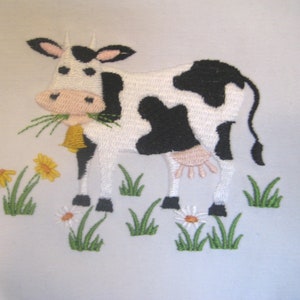 May include: Embroidered design of a black and white cow with a pink nose, eating green grass. The cow is standing in a field of green grass with yellow flowers and white daisies.