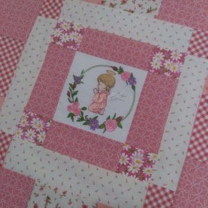 May include: A pink and white patchwork quilt with a floral design and an embroidered image of a praying angel in a wreath of flowers. The quilt has a gingham pattern in some of the squares.