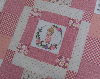 Puede incluir: Una colcha de retazos rosa y blanca con un diseño floral y una imagen bordada de un ángel rezando en una corona de flores. La colcha tiene un patrón de cuadros en algunos de los cuadrados.