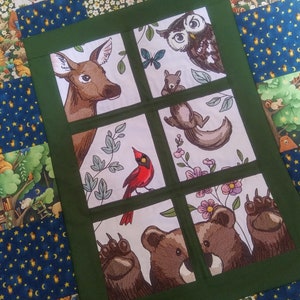 May include: A green fabric panel with a white background featuring embroidered images of a deer, owl, squirrel, cardinal, bear, and paw print. The images are arranged in a grid pattern within a green frame.