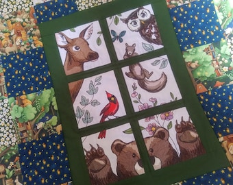 Puede incluir: Un panel de tela verde con un fondo blanco que presenta imágenes bordadas de un ciervo, un búho, una ardilla, un cardenal, un oso y una huella de pata. Las imágenes están dispuestas en una cuadrícula dentro de un marco verde.