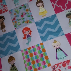 May include: A white quilt with a patchwork design featuring embroidered images of princesses and mermaids. The quilt has a variety of patterns including pink and blue chevron, polka dots, and floral designs.