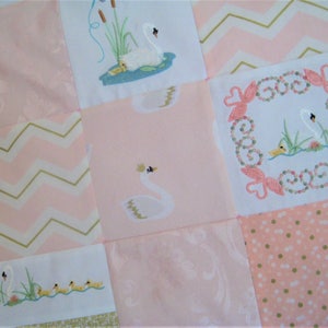May include: A pink and white patchwork quilt with embroidered swans and ducklings. The quilt is made of different fabrics with various patterns, including polka dots, chevron, and floral.