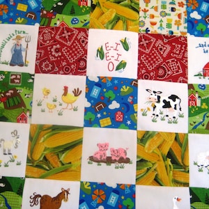 May include: A colorful patchwork quilt with various farm-themed squares. The squares feature images of a cow, pig, corn, a barn, and a goat. The quilt is made of white fabric with colorful embroidered designs.